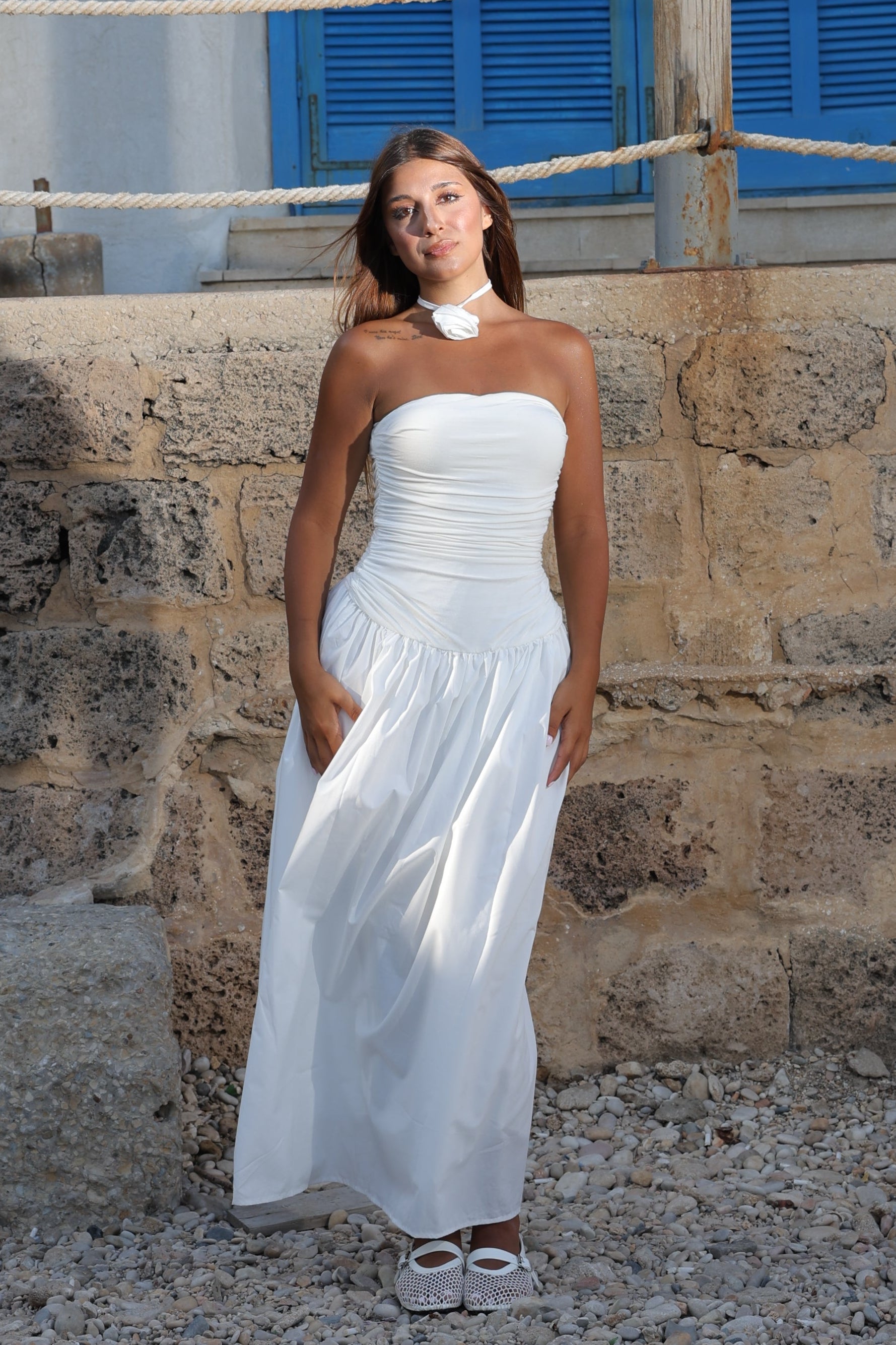 Woman in a white dress standing against a stone wall with blue shutters.